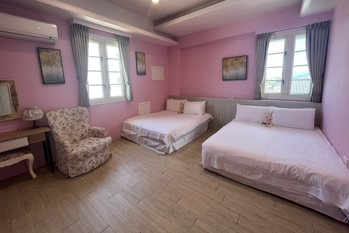 A pink-themed hotel room with two single beds, a chair, a desk, and a window with curtains.