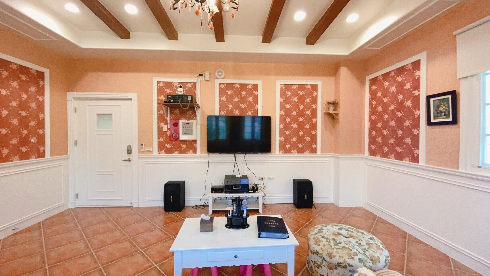 A spacious room with a large flat-screen TV, a white table with electronic devices, and a floral-patterned ottoman.