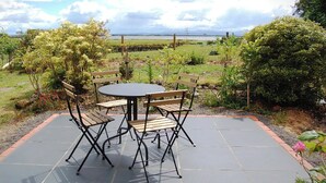 Un patio al aire libre con una mesa redonda y cuatro sillas, rodeado de un jardín con diversas plantas y una vista a un cuerpo de agua al fondo.