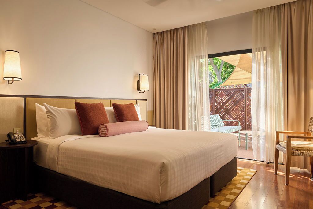 A hotel room with a large bed, two bedside lamps, a telephone, and a view of an outdoor patio with a chair and a table.