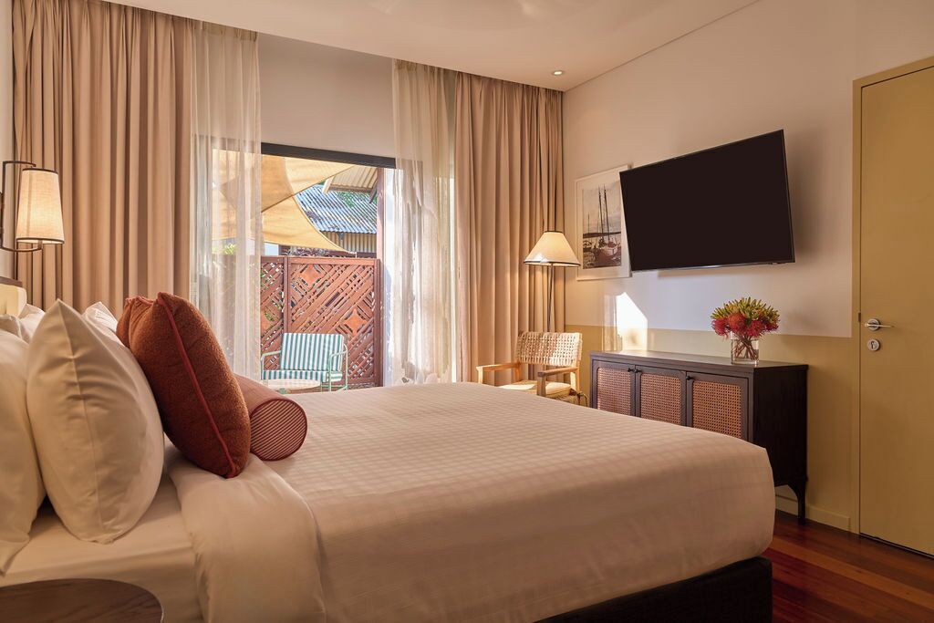 A hotel room with a large bed, a flat-screen TV on the wall, a desk with a lamp, and a view of a wooden deck through the window.