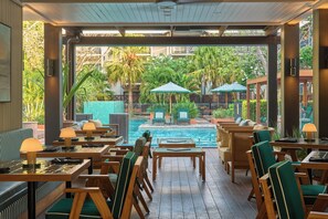 Zona junto a la piscina con mesas y sillas de madera, sombrillas y vistas a la piscina y al follaje.
