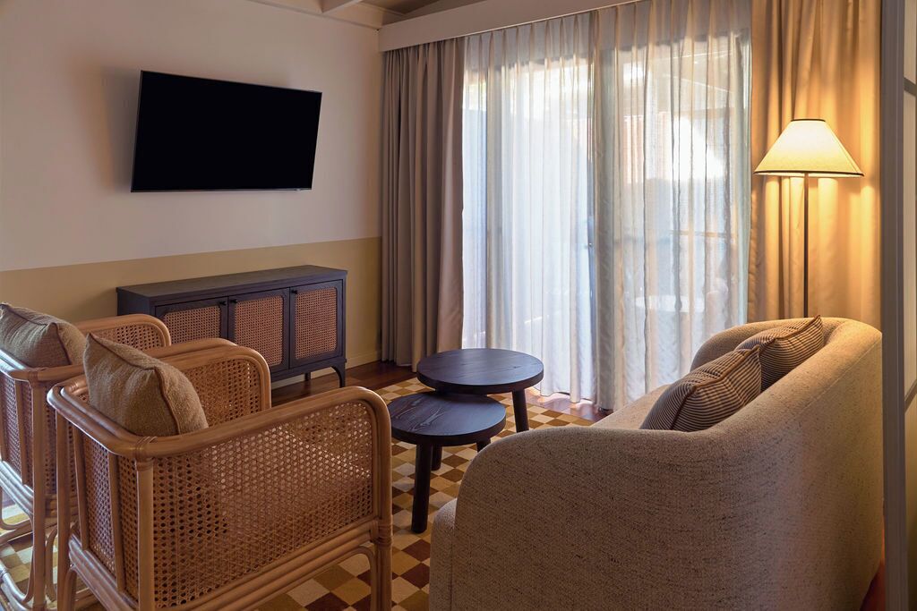 Et hotelværelse med et fladskærms-tv på væggen, en fletsofa, et rundt bord og en lampe.