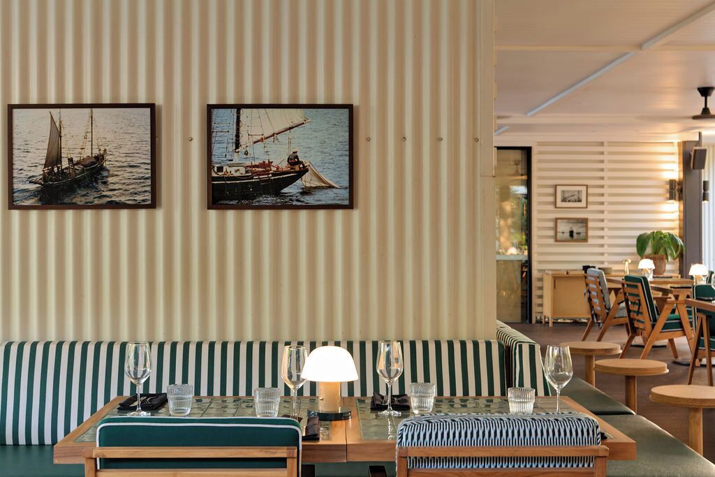 Een eethoek met gestreepte wanden, een tafel gedekt met glazen en een lamp, en twee ingelijste scheepvaartschilderijen.