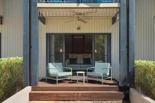 Un espacio al aire libre techado con dos sillas a rayas y una mesa pequeña, un ventilador de techo y un ventanal corredera que da a una habitación con un sofá y cojines.