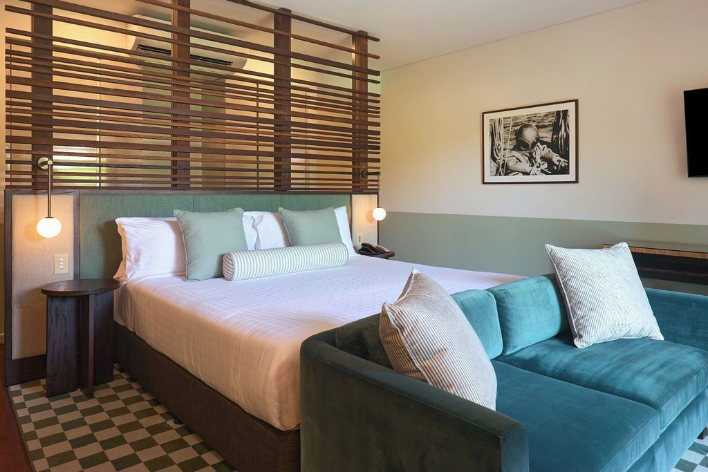 Une chambre d’hôtel avec un lit, un canapé, un sol à carreaux, une tête de lit en bois et un tableau encadré au mur.