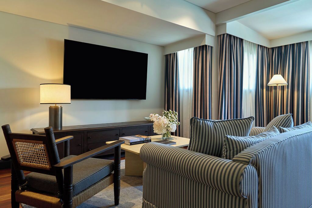 A hotel room with a large flat-screen TV mounted on the wall, a striped sofa, a desk with a lamp, and a chair.