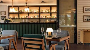 Una zona de bar con mostrador de madera, estantes llenos de botellas y luces colgantes. Hay dos mesas con sillas, una de ellas con un sillón tapizado en verde.