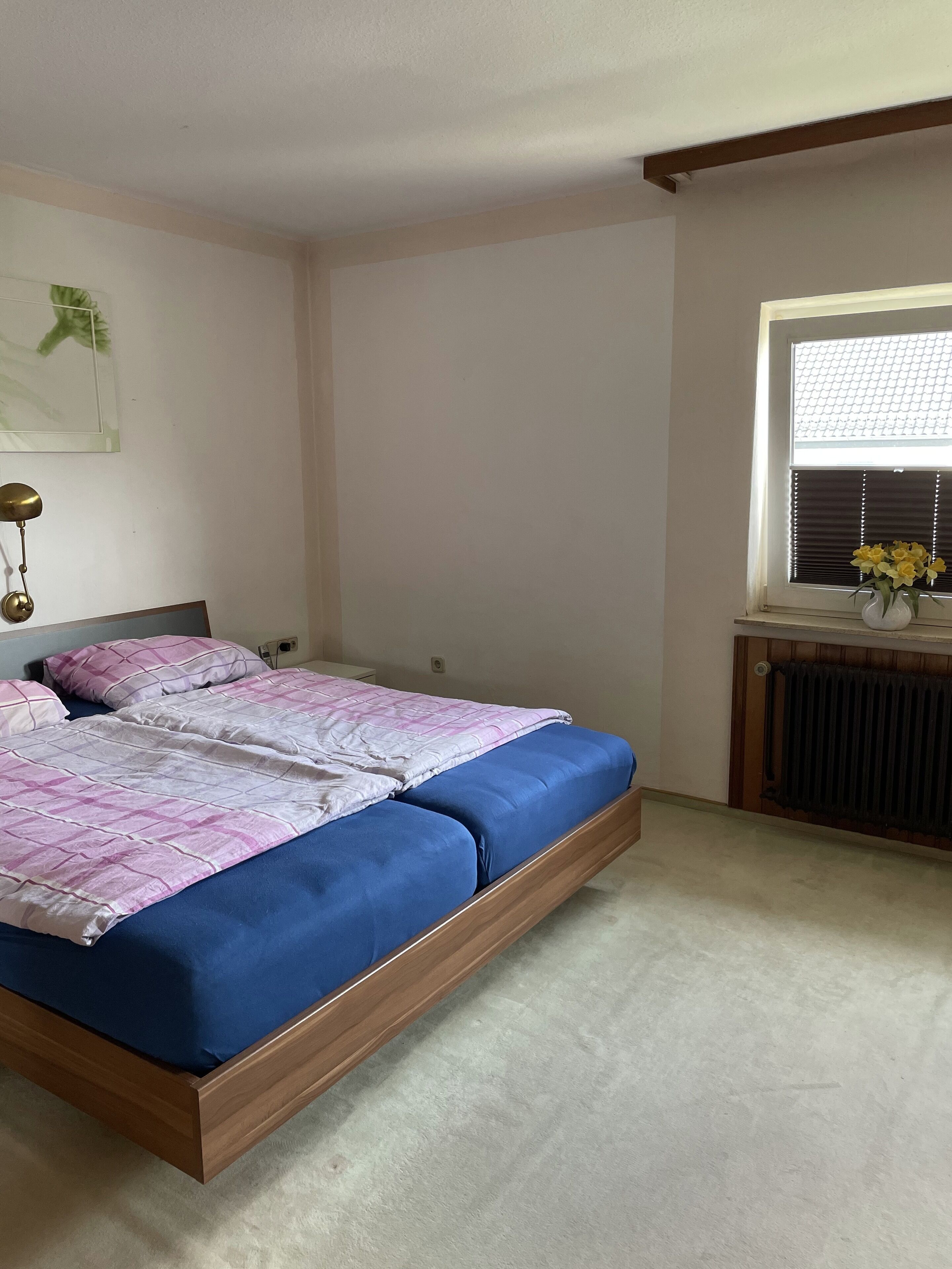 Ein Schlafzimmer mit Doppelbett, Heizkörper, Fenster mit Jalousien und einer Wandlampe.
