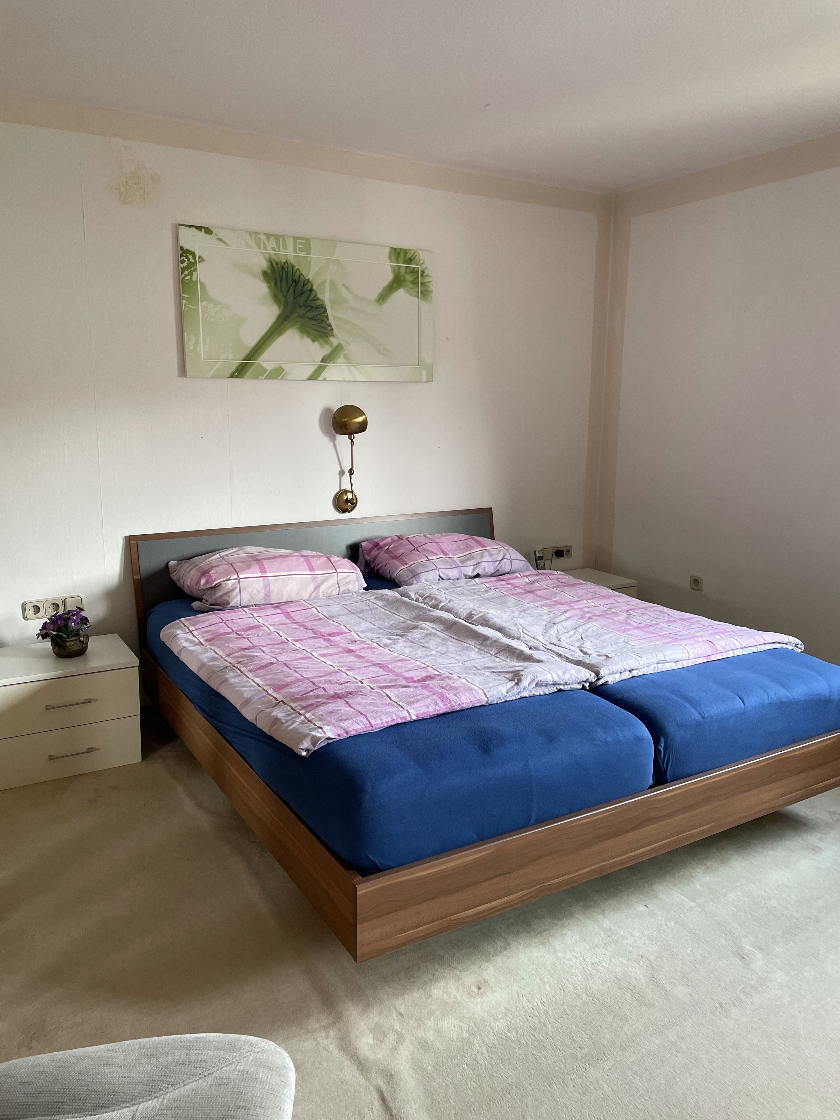 Een slaapkamer met een bed, nachtkastje, een wandlamp en een lijst met kunstwerk.