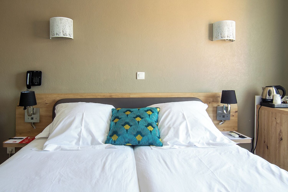 A hotel room with a bed, two wall-mounted lamps, a telephone, and a kettle on a cabinet.