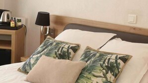 A neatly made bed with two pillows, one plain and one with a tropical leaf pattern, in a room with a wooden headboard and a nightstand with a lamp. - Azur Hotel (Balaruc-les-Bains)