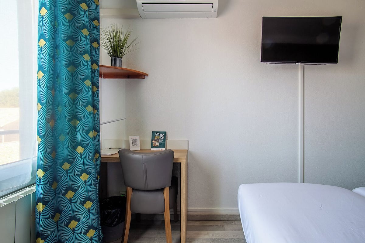 A hotel room with a desk, chair, and a bed. There is a TV mounted on the wall and a plant on a shelf.