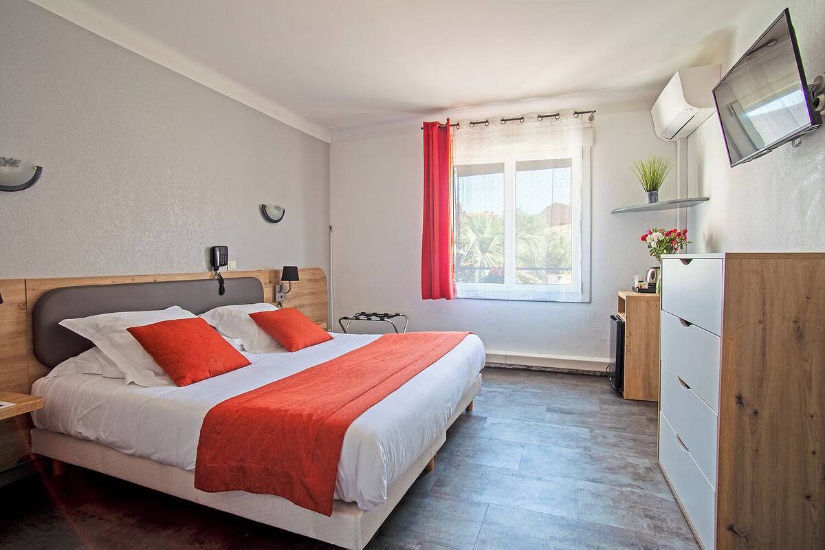 A hotel room with a bed, a nightstand, a desk, a chair, a TV, and a window with curtains.