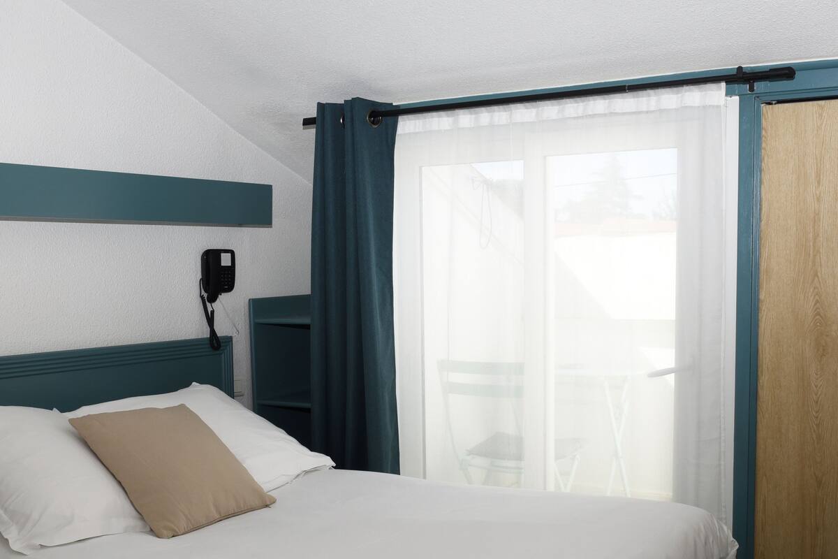 A hotel room with a bed, a bedside table with cups and a tray, a phone on the wall, and a window with curtains.