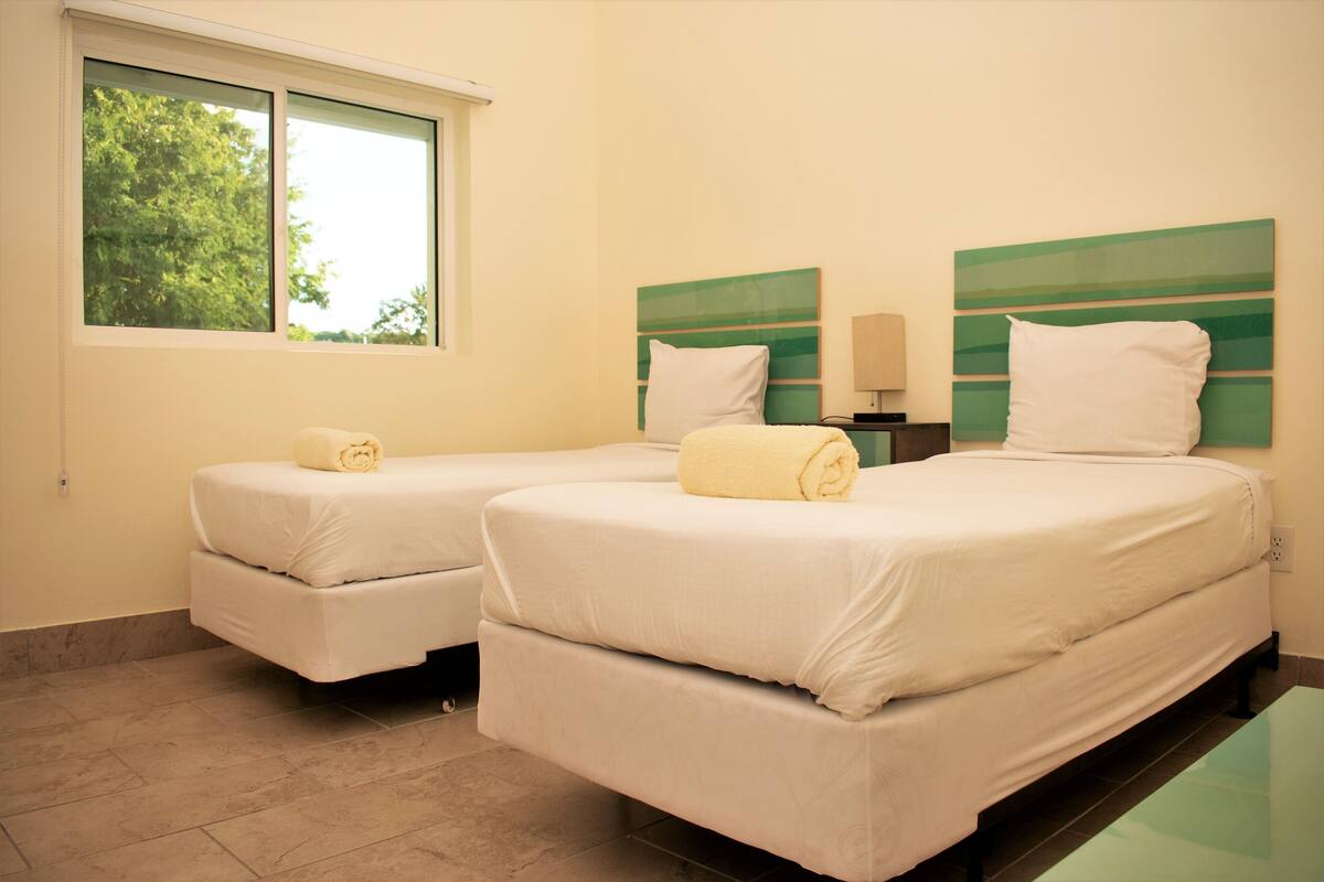 A hotel room with two beds, each with a white bedspread and a rolled towel. There is a window with a view of trees, and the room has a tiled floor.