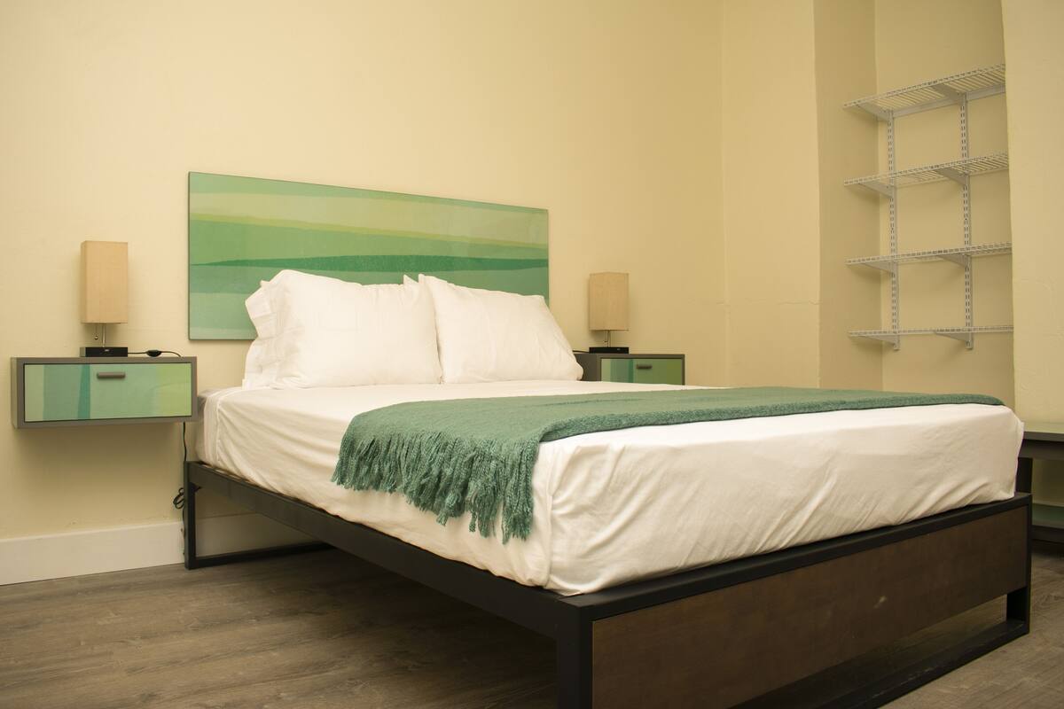 A neatly made bed with white linens and a green throw, flanked by two bedside tables with lamps, in a room with a green abstract painting on the wall and a metal shelf unit.