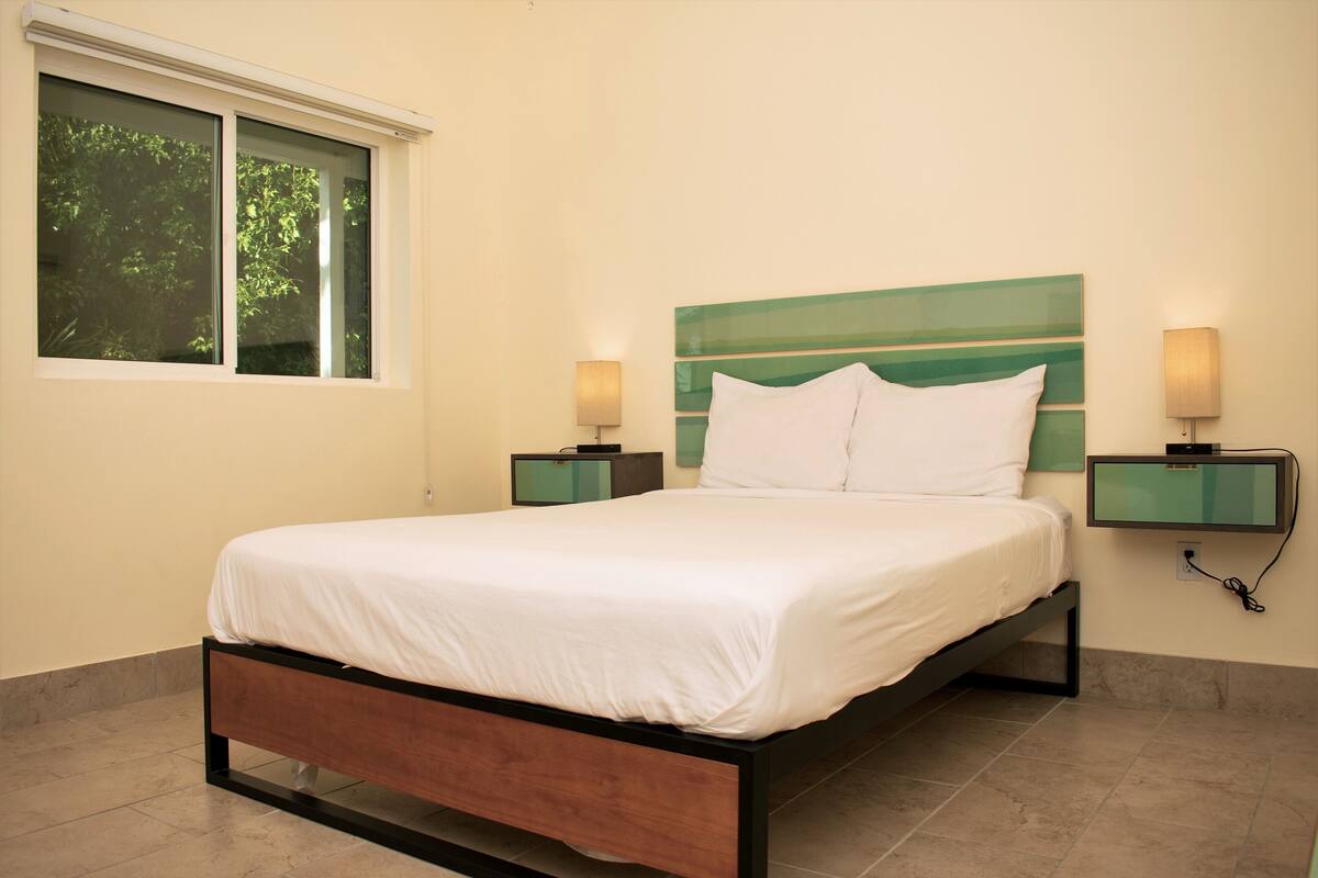 A neatly made bed in a room with a large window, two bedside tables, and a green headboard.