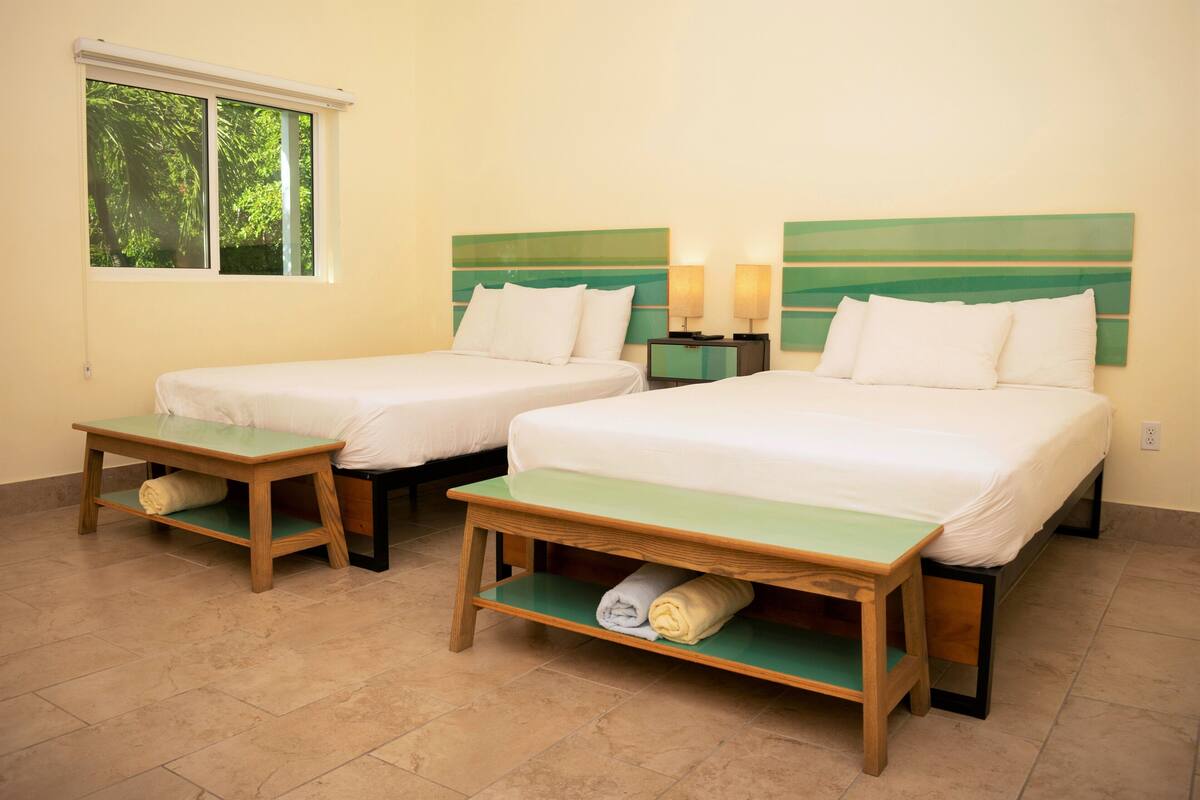 A hotel room with two single beds, a small table between them, a nightstand with a lamp, and a window with a view of trees.