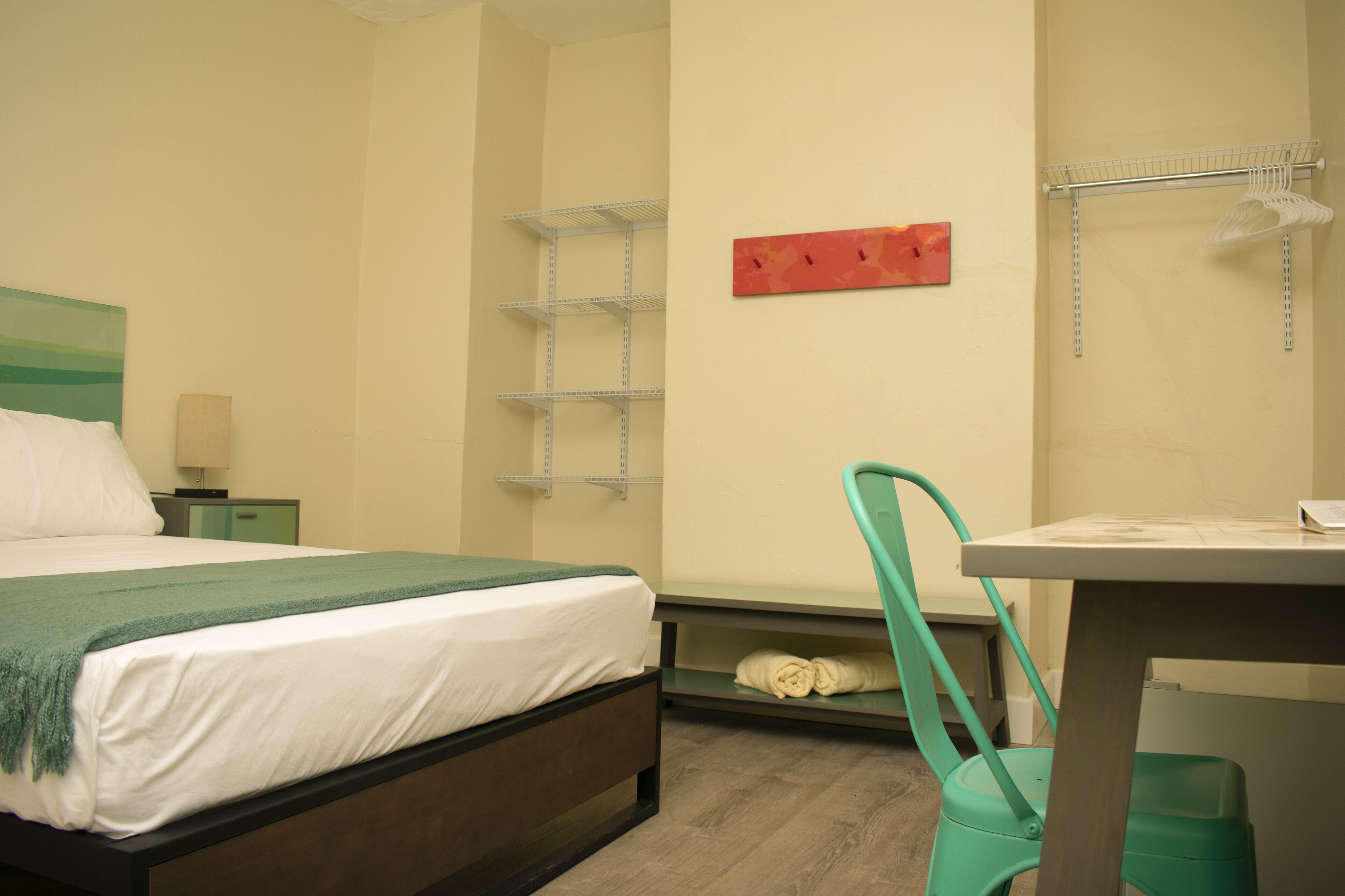 A hotel room with a bed, a desk, a chair, a shelf, and a wall-mounted towel rack.