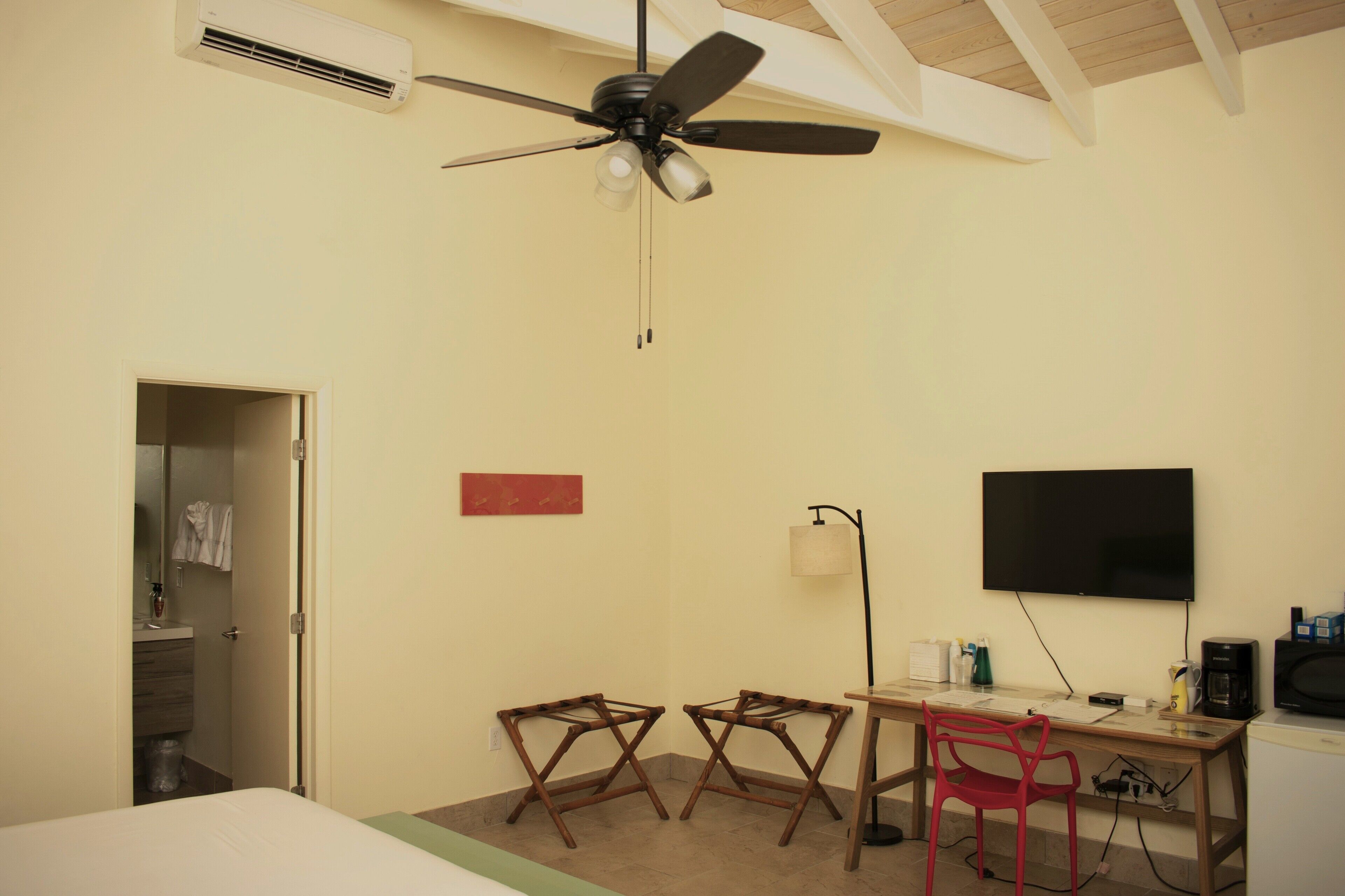 A hotel room with a bed, a desk with a chair, a TV, a ceiling fan, and a door leading to a bathroom.