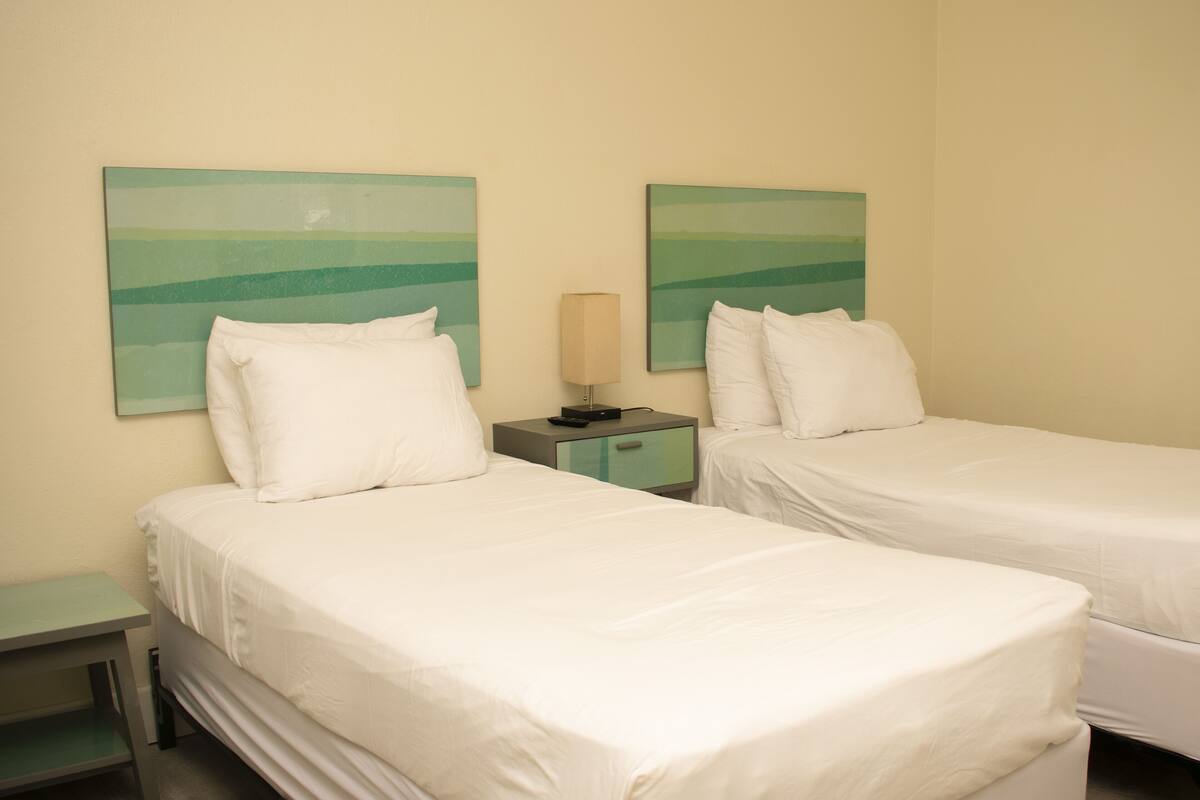 A hotel room with two single beds, a bedside table with a lamp, and abstract wall art.