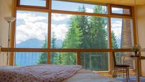 A bedroom with a large window, a bed, a chair, and a desk with a plant.