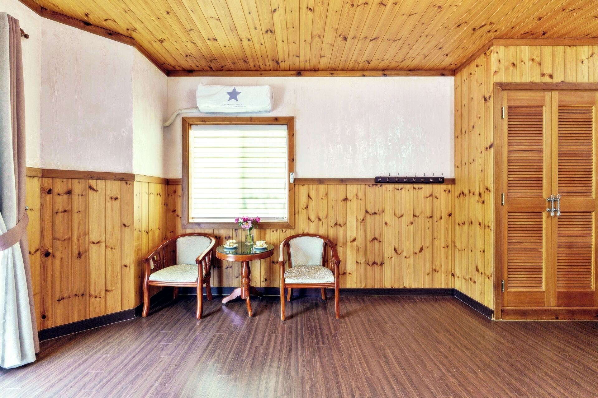 A room with wooden walls and ceiling, a wooden door, a window with blinds, two chairs, a small table with a vase of flowers, and a star-shaped decoration on the wall.