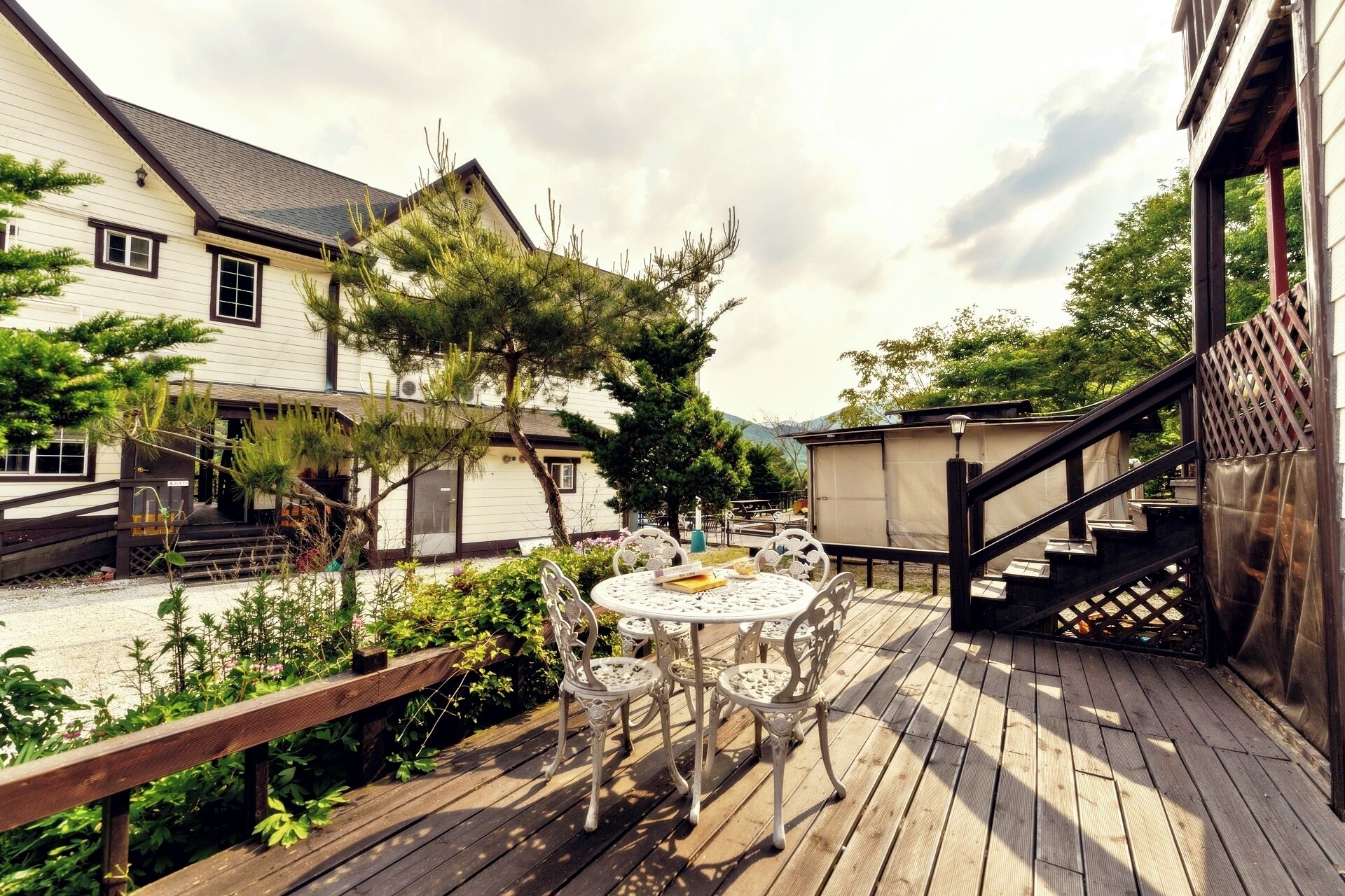 A wooden deck with a round table and chairs, surrounded by greenery and a building with a staircase.