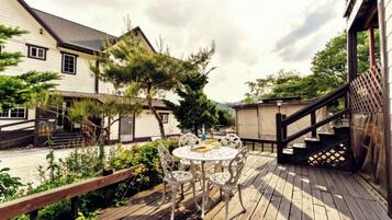 A wooden deck with a round table and chairs, surrounded by greenery and a building with a staircase.