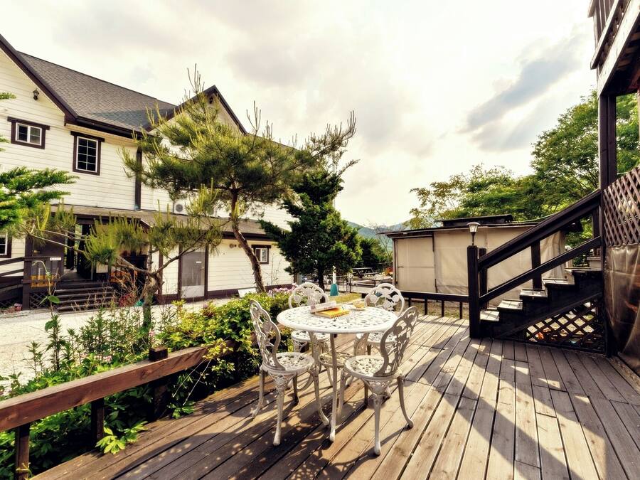 A wooden deck with a round table and chairs, surrounded by greenery and a building with a staircase.
