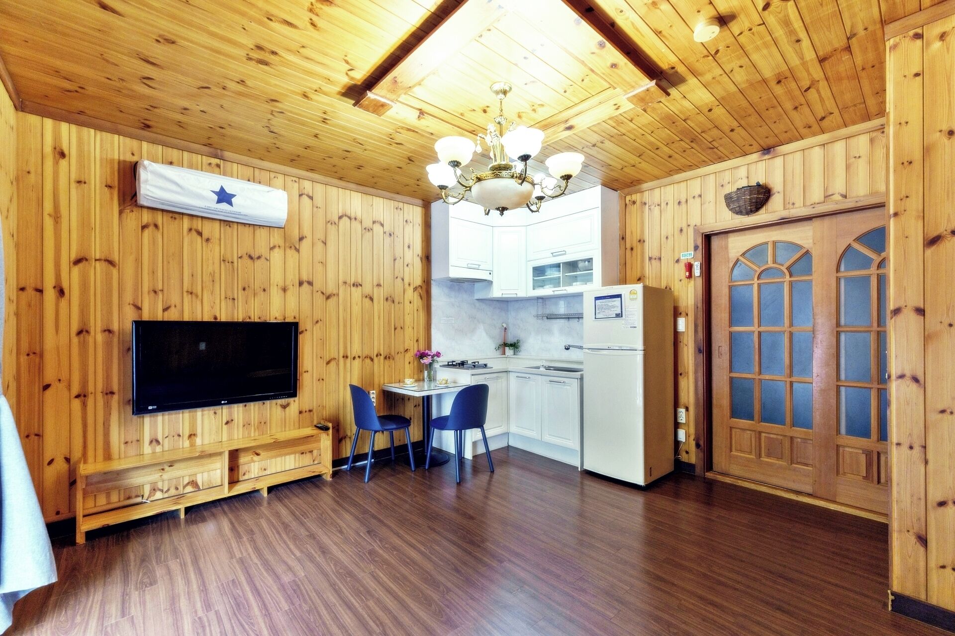 Interior em madeira com televisão, área de refeições, cozinha e uma grande porta de madeira.
