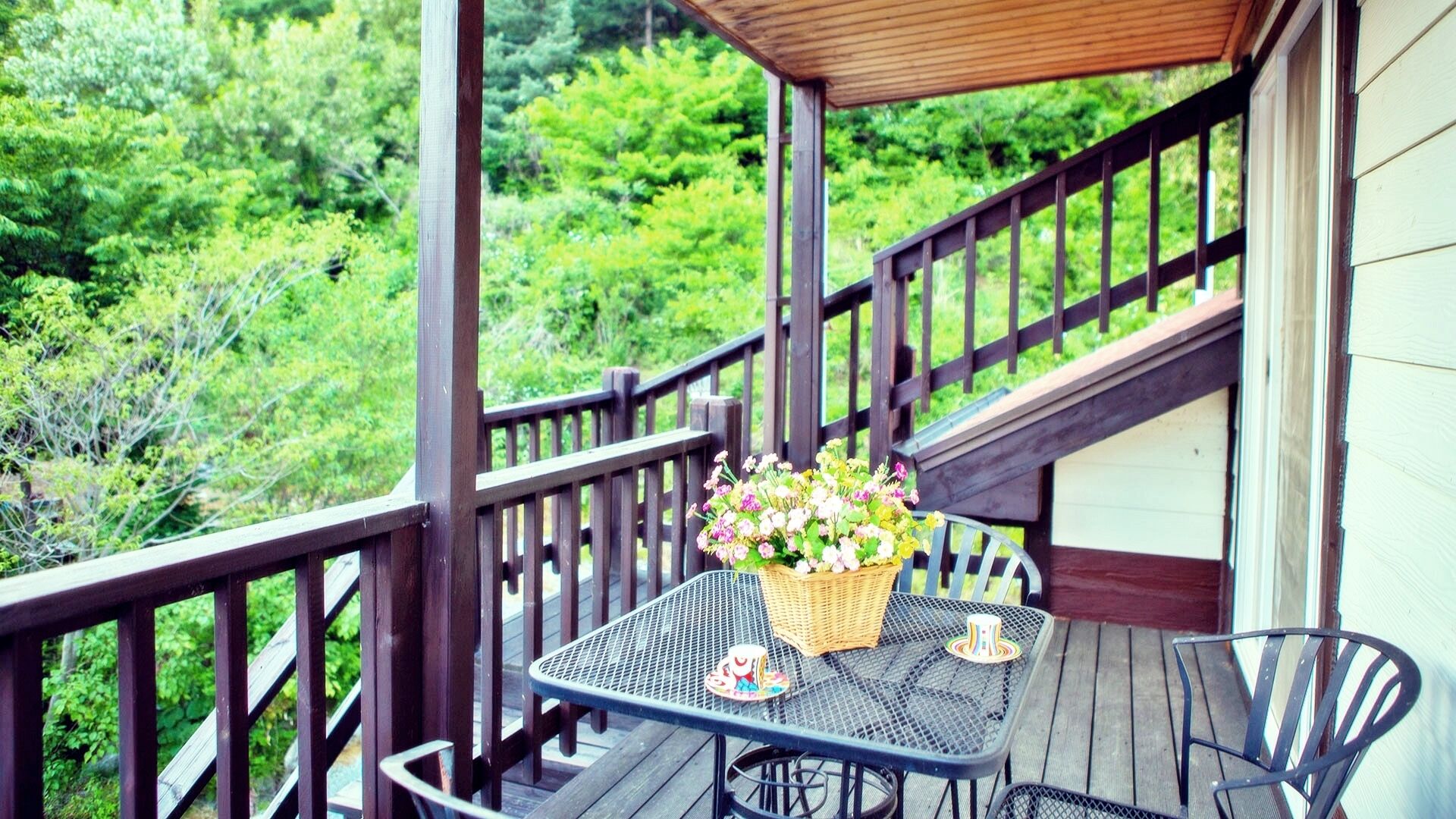 A wooden deck with a table and chairs, a basket of flowers, and a view of green foliage.