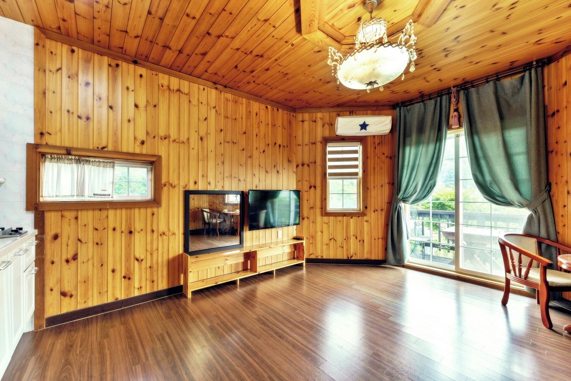 A wooden-paneled room with a television, a dining table, and a large window with curtains.