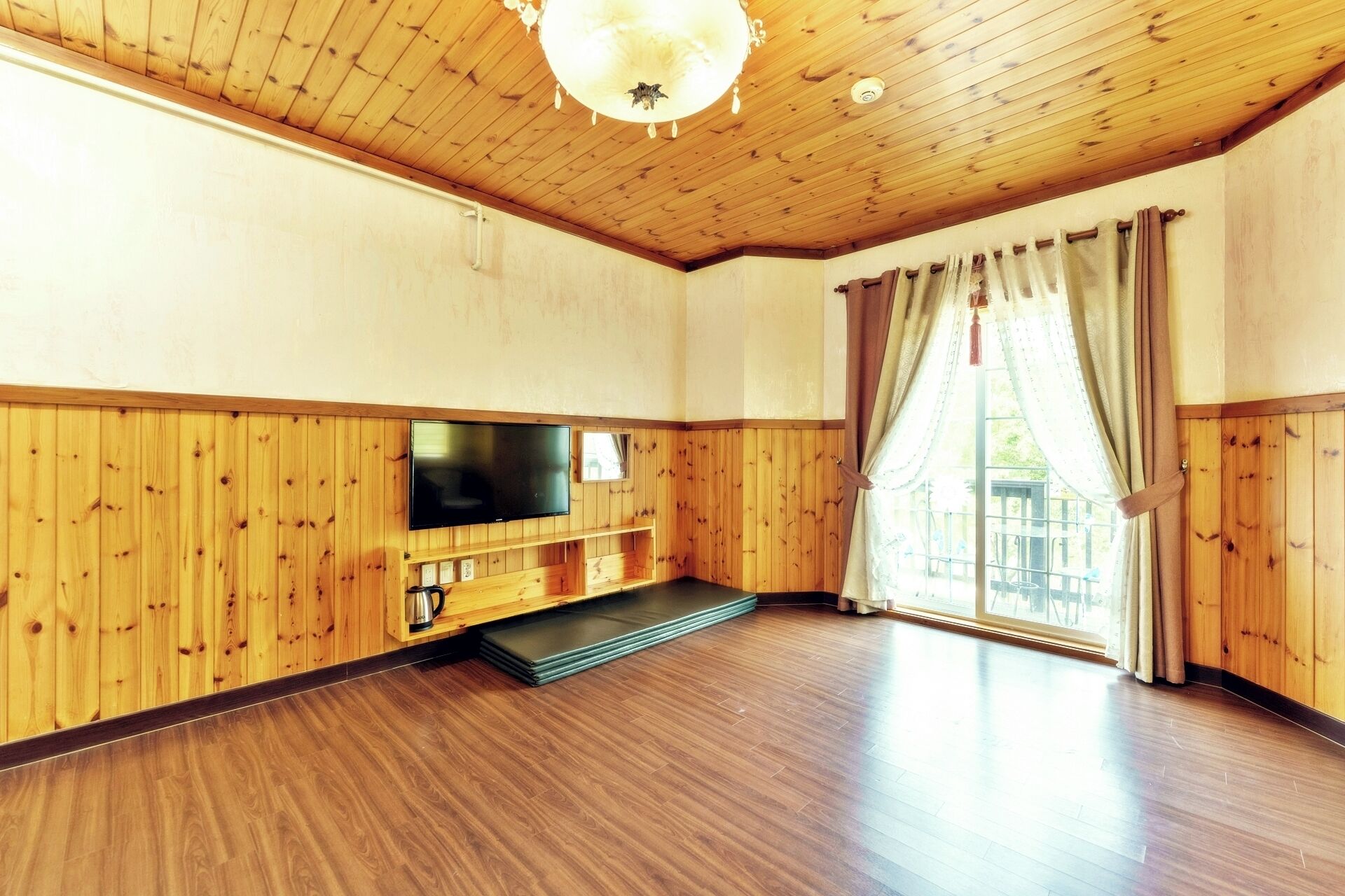 A spacious room with wooden paneling, a flat-screen TV, a coffee table, and a large window with curtains.