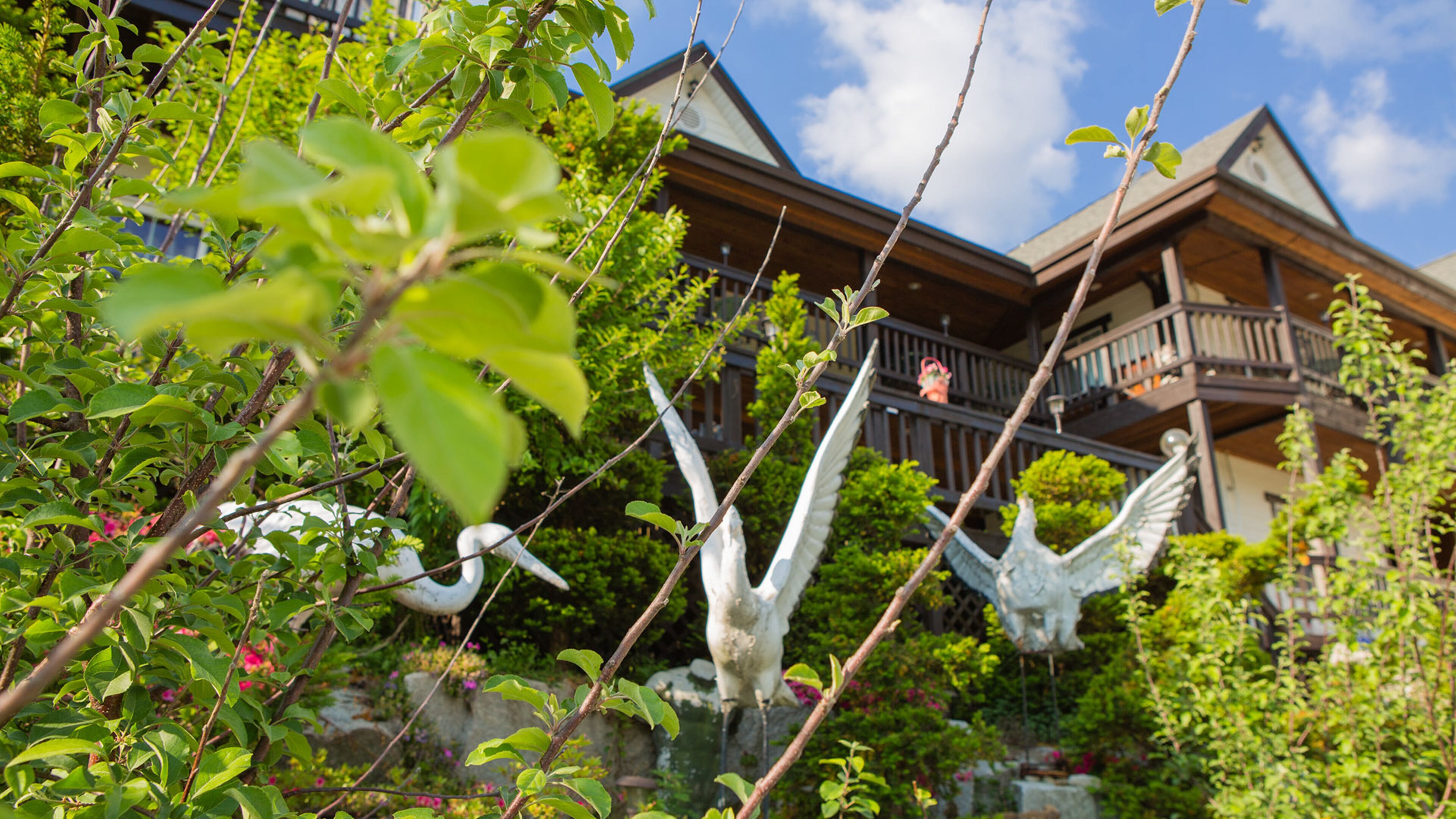 Una casa con balcón, rodeada de abundante vegetación y esculturas decorativas de aves.