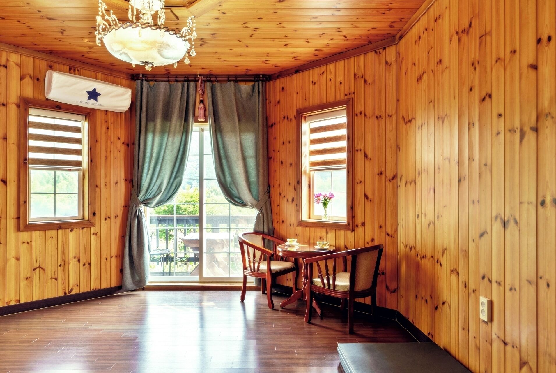 A wooden-paneled room with a dining area, a chandelier, and a view through a large window.