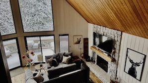 A cozy living room with a fireplace, large windows, and a stone wall. - SPACIOUS BLUE JAY CABIN WITH VAULTED CEILINGS! (Lake Arrowhead)