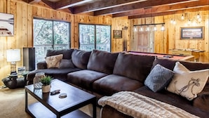 A cozy living room with a dark brown leather sofa, a coffee table with decorative items, and a view of the outdoors through large windows. - SPACIOUS BLUE JAY CABIN WITH VAULTED CEILINGS! (Lake Arrowhead)