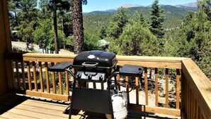 A deck with a barbecue grill and a water dispenser, overlooking a forested area with mountains in the background. - Mountain Majesty (Ruidoso)
