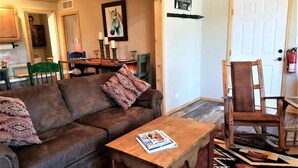 A cozy living room with a brown leather sofa, a wooden coffee table, and a patterned rug. A wooden chair is placed near a door with a fire extinguisher visible. - Mountain Majesty (Ruidoso)