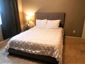 Una cama bien hecha con un cubrecolcha acolchado, una lámpara de noche y una cortina estampada.