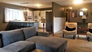 A living room with a gray sectional sofa, a white armchair, and a small table with a lamp. There is a kitchen in the background with white cabinets and a dining area with a table and chairs. - Friendly Nabors (Ruidoso)