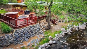 Une terrasse en bois avec un jacuzzi et des meubles d’extérieur, entourée d’un mur de pierres et bordée d’un petit ruisseau.