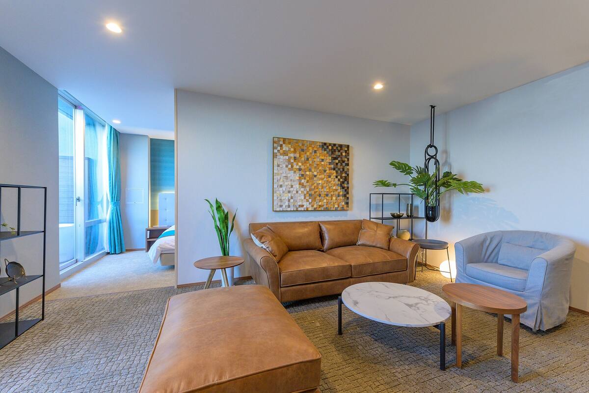 A modern living room with a brown sofa, a light grey armchair, a coffee table, and a small side table. There is a large abstract painting on the wall, a potted plant, and a window with curtains.