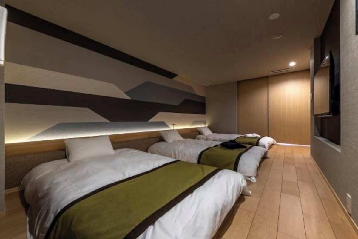 A hotel room with three beds arranged in a row, a flat-screen TV mounted on the wall, and a modern geometric wall design.