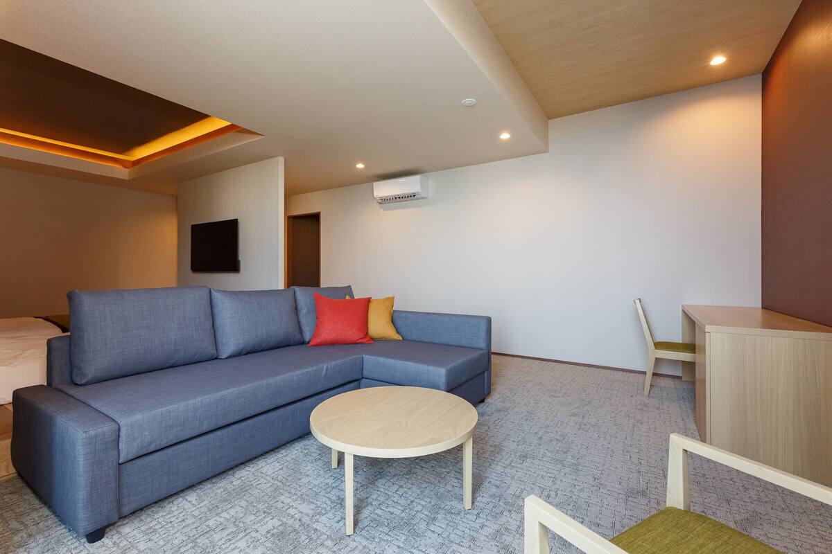 A modern living room with a sectional sofa, a round coffee table, a wooden desk, and a flat-screen TV mounted on the wall.