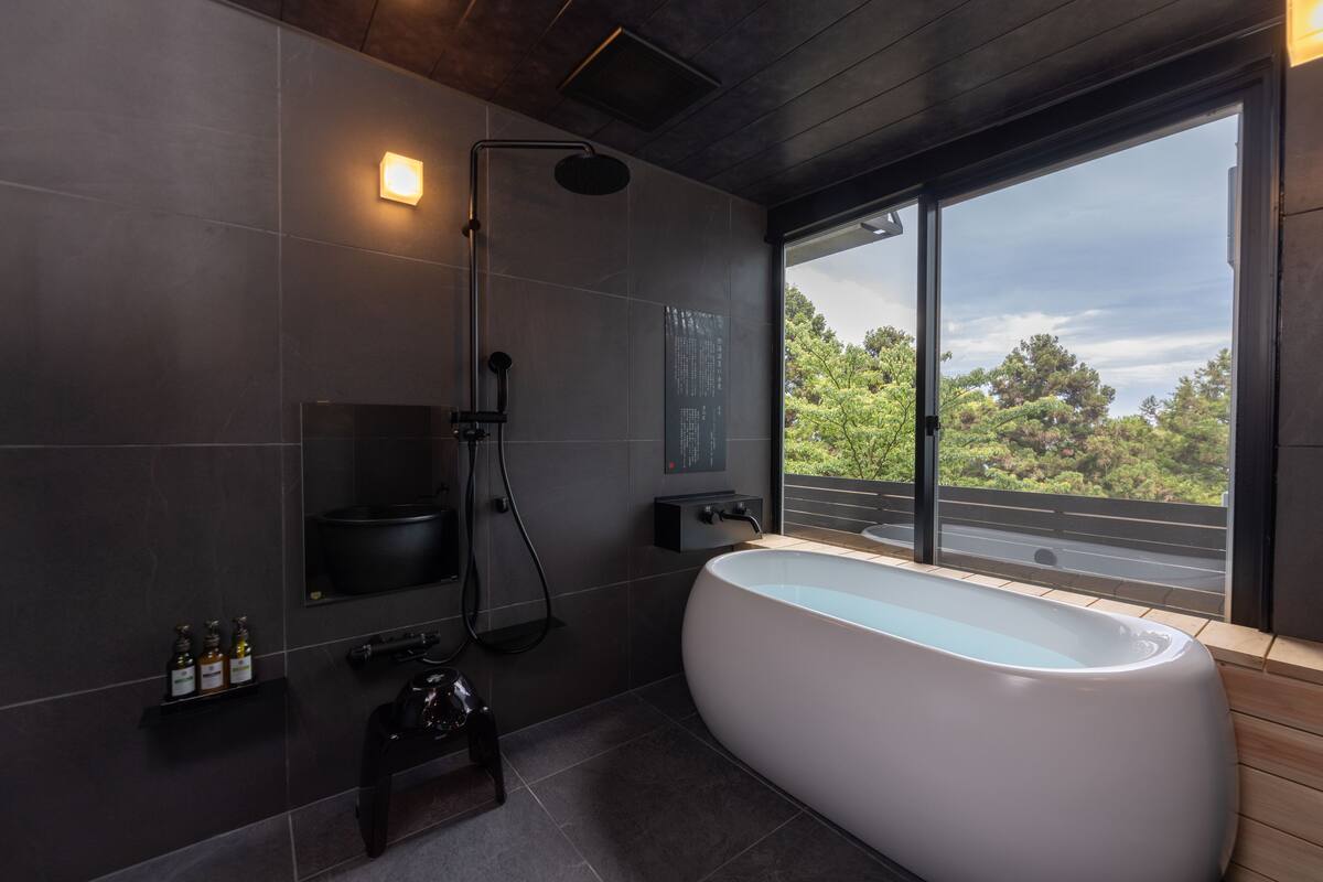 A modern bathroom with a freestanding bathtub, a showerhead, and a large window offering a view of greenery.