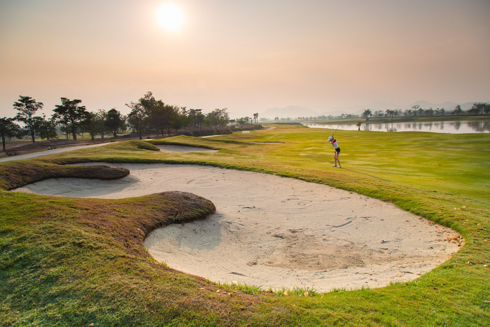 A golf course with sand bunkers, a water body, and trees in the background.