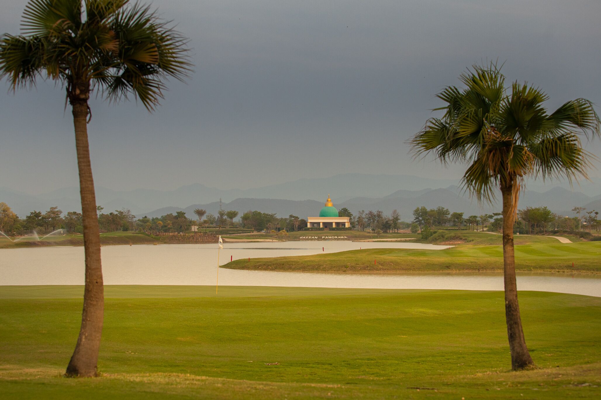 A golf course with a building that has a green dome roof, surrounded by palm trees and a body of water.